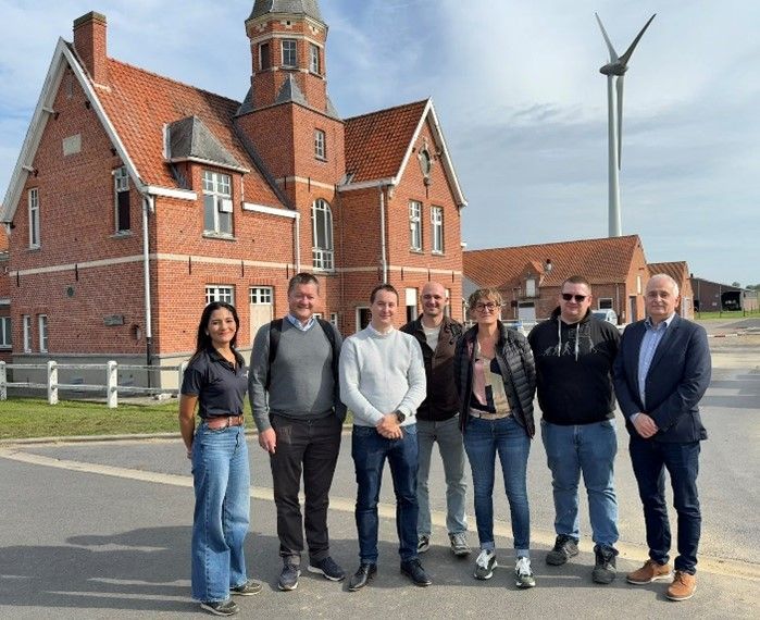 A group of eight people stand in front of a red brick building with a tower and a wind turbine in the background.