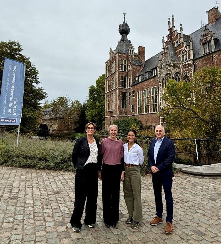 Four people stand outside a historic castle with a tall spire and red-brown brick facade.