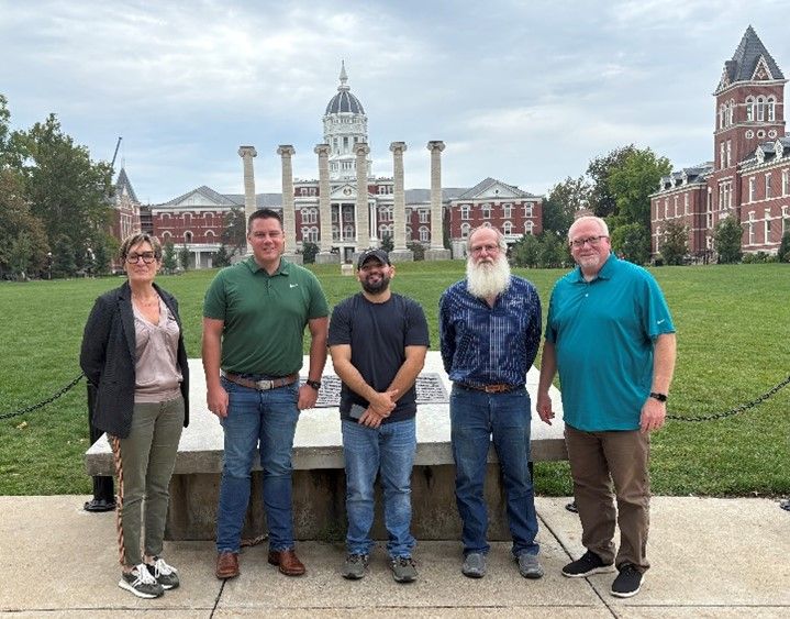 Five people stand in front of a large, ornate building with a dome and clock tower on a grassy lawn.