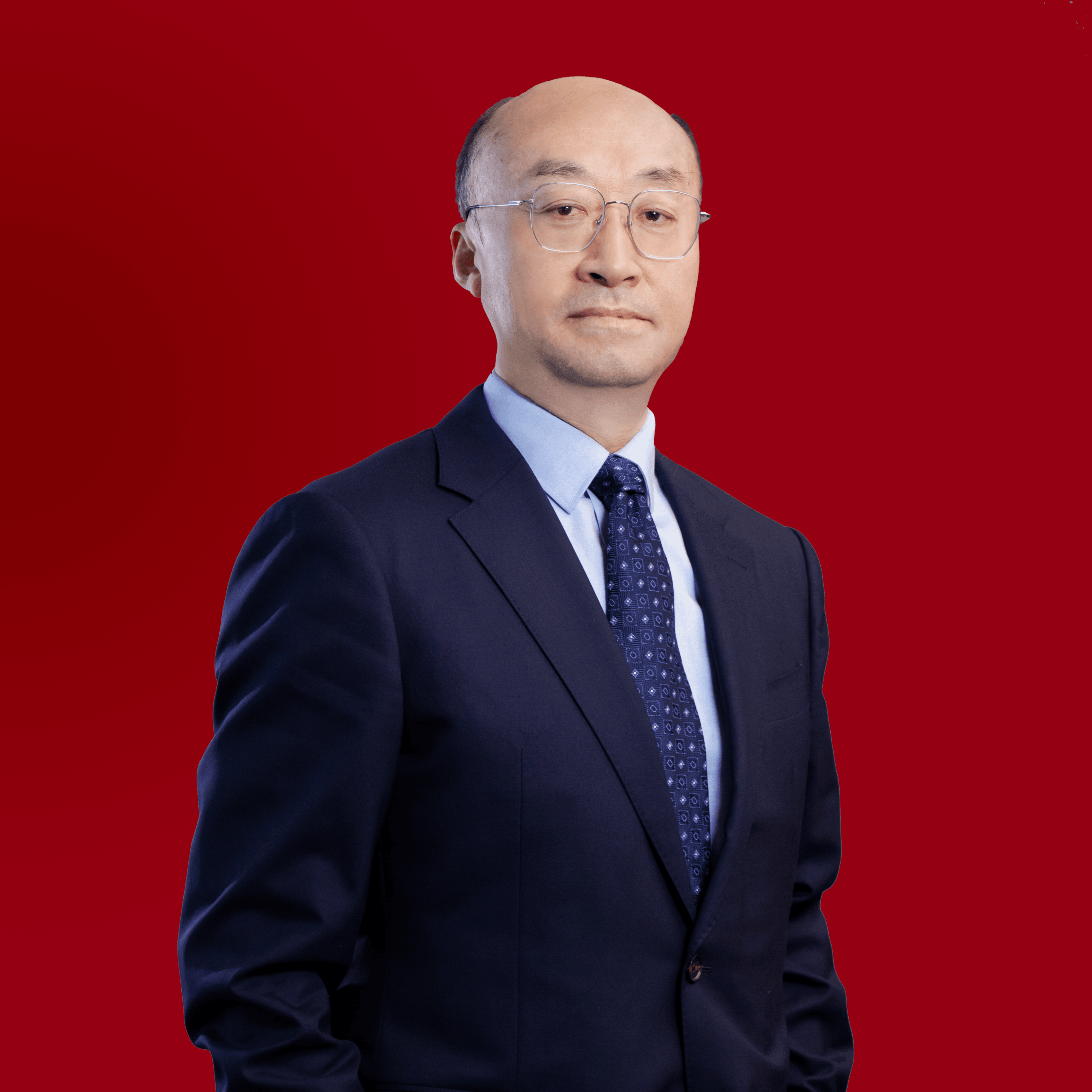 Un homme distingué, vêtu d'un costume bleu foncé et portant des lunettes, se tient debout devant un fond rouge.